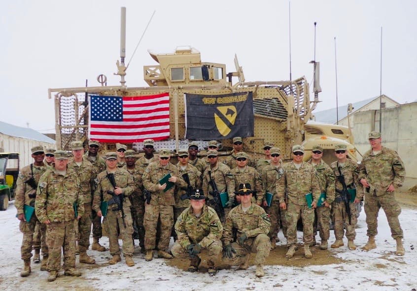 A 1st Cavalry Division platoon formed up in the snow beneath the U.S. flag and the 1st Cavalry guidon, an MRAP behind them, after a unit ceremony downrange
