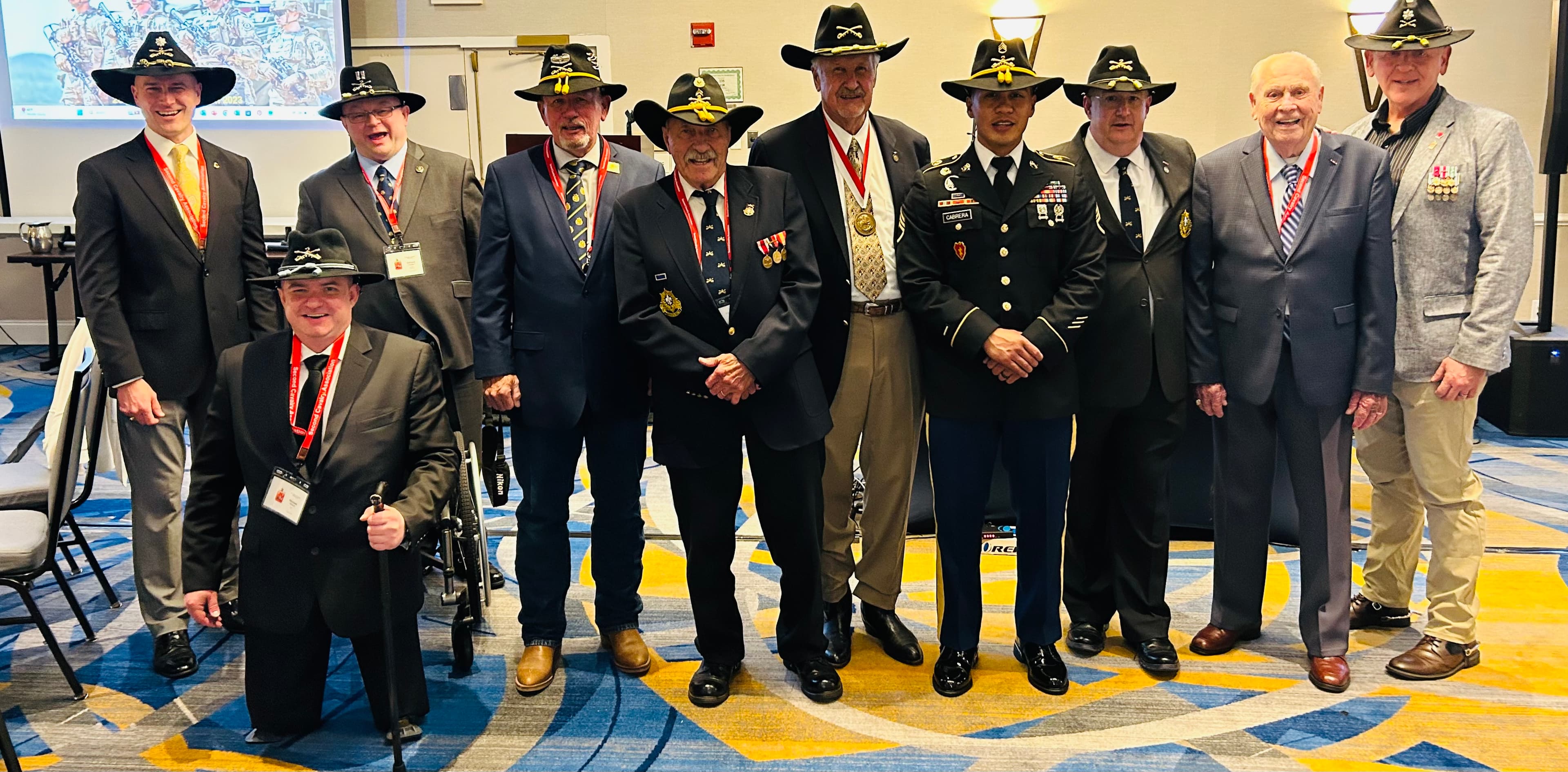 Veterans gathered at a unit association reunion, multi-generational cavalry troopers in Stetson hats alongside a serving Soldier in uniform.